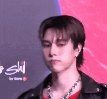 a young man wearing a leather jacket and a necklace is standing in front of a pink background .