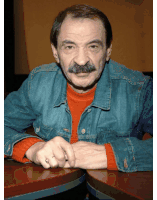 a man with a mustache is sitting at a table with his hands folded