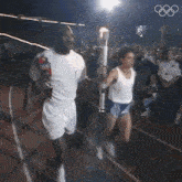 a man and a woman carrying a torch on a track with the olympics logo in the background