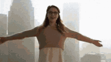 a woman wearing glasses is standing in front of a city skyline with her arms outstretched .