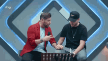two men are sitting next to each other and one is pointing at the other 's hand .