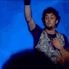 a man wearing a purple shirt and a paisley vest is smiling and raising his arm in the air .