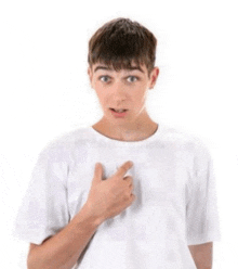 a young man in a white t-shirt is pointing at his chest with his finger .