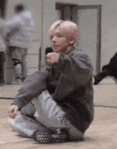 a young man with pink hair is sitting on the floor and pointing at the camera