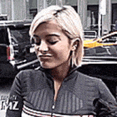 a woman with blonde hair is wearing a black jacket and standing in front of a car .