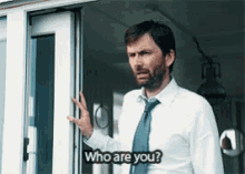 a man in a white shirt and tie is standing in front of a door asking who are you .