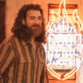 a man with long hair and a beard is smiling in front of a neon sign .
