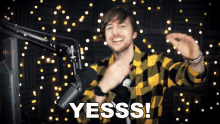 a man in a yellow and black plaid shirt stands in front of a microphone with the words yesss written below him