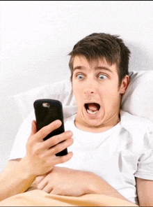 a man laying in bed looking at a cell phone with his mouth open