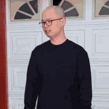 a bald man wearing glasses and a black sweater is standing in front of a garage door