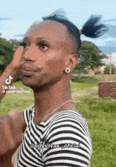 a man wearing a striped shirt and earrings is standing in a field with his hair in a ponytail .