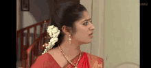 a woman wearing a red saree and a gold necklace is standing in front of a staircase .