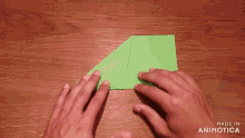 a person folding a piece of green paper on a wooden table with the words made in animatica on the bottom