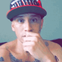 a shirtless man wearing a hat and a tattoo on his chest is covering his mouth with his hand .