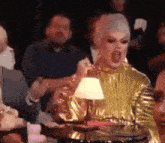 a woman in a gold dress is sitting at a table with a lamp and shouting .