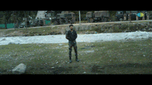 a man in military uniform stands in front of a river with military vehicles in the background
