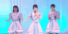 three girls are singing into microphones on a stage and one of them is wearing a blue and white dress