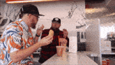 two men are sitting at a counter eating sandwiches and drinking orange juice from a cup that says ' donut shop '