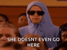 a woman wearing sunglasses and a hooded jacket is sitting in a crowd of people and says she does nt even go here