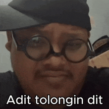 a man wearing glasses and a hat says " adit tolongin dit " on the bottom