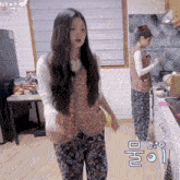 a woman in a floral vest stands in a kitchen while another woman prepares food