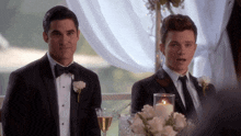 two men in tuxedos are sitting at a table with a candle and a glass of champagne