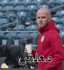 a bald man in a red hoodie is holding a cup in front of a stadium .