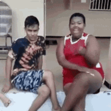 a man and a woman are sitting on a bed . the woman is wearing a red tank top .