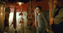 a man in a grey sweater stands in front of meat hanging from hooks