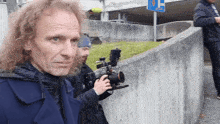 a man with curly hair is holding a camera in front of a sign that says ' a ' on it
