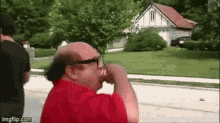 a bald man in a red shirt and sunglasses is drinking from a cup .