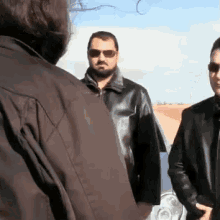 a man wearing sunglasses and a leather jacket stands next to two other men