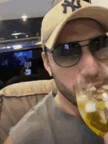 a man wearing a ny hat and sunglasses drinks a glass of beer