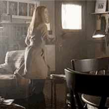 a woman in a trench coat is standing in a living room looking out a window .