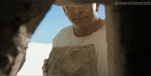 a man in a white shirt is looking through a hole in a rock with the hashtag @itsscottiesstark on the bottom
