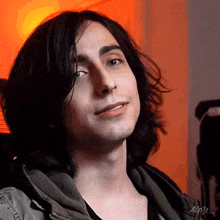 a young man with long black hair is sitting in front of an orange wall .