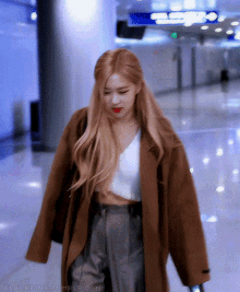 a woman wearing a brown coat and a white crop top is walking through an airport