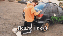 a man in an orange hoodie is kneeling next to a black car with graffiti on the back