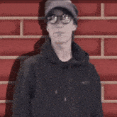 a person wearing glasses and a hat stands in front of a brick wall