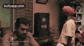 a man in a turban is talking on a cell phone while another man stands behind him .