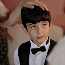 a young boy in a tuxedo and bow tie is being touched by a woman 's hand