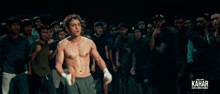a shirtless man is standing in front of a group of people with kahar written on the bottom