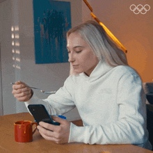 a woman sitting at a table looking at her phone and eating cereal