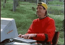 a man in a red shirt is typing on a keyboard in front of a computer .