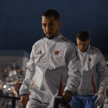 a man in a white adidas jacket walks with another man in a blue shorts