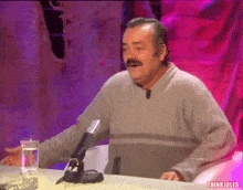 a man with a mustache is sitting at a table with a microphone and a glass of water in front of him