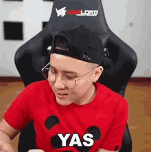 a man wearing glasses and a baseball cap is sitting in a chair with the word yas on his shirt .