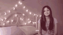 a young girl is sitting on a bed in front of a bed with a string of lights hanging from the headboard .