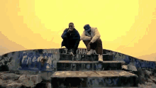two men sit on a set of stairs with graffiti on the wall that says " t.a. chaka "