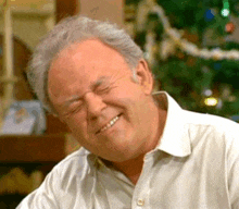 an older man in a white shirt is laughing with his eyes closed in front of a christmas tree .